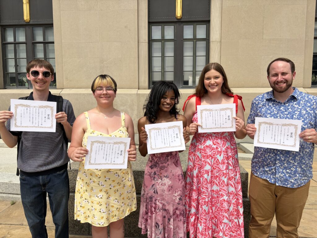 Five MTSU students stand outdoors in front of a stone building, smiling and holding certificates with Japanese writing after receiving awards at a Japanese language contest.