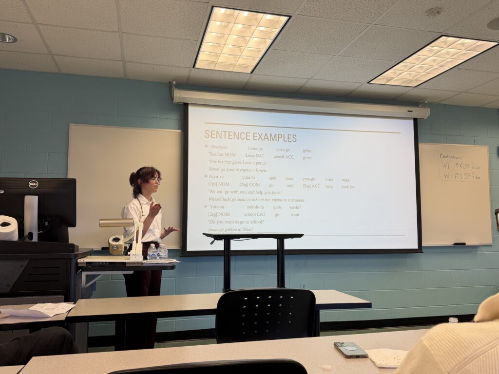 A student presenter stands at the front of a classroom explaining example sentences in Japanese, with a slide displaying grammatical structures and translations projected behind them.