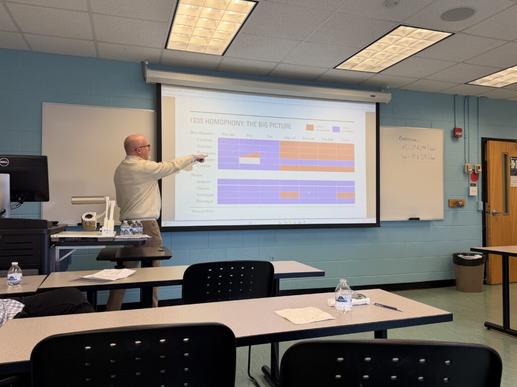 A faculty presenter gestures toward a projected slide showing a chart on homophony across Romance languages, including Castilian, Galician, Portuguese, and others, during a classroom presentation.
