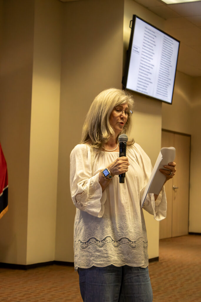 Dr, McRae stands at the front of a room reading from a paper into a microphone during a poetry event, with a screen displaying text behind her.
