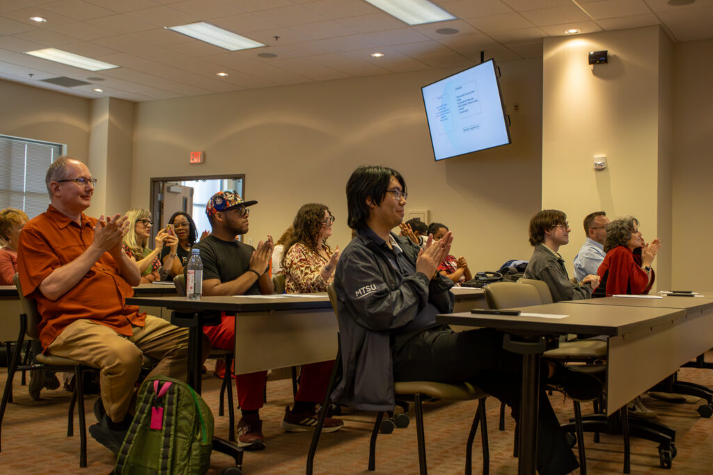 An audience seated in a classroom applauds during a WLLC event, with attendees smiling and facing the front of the room.