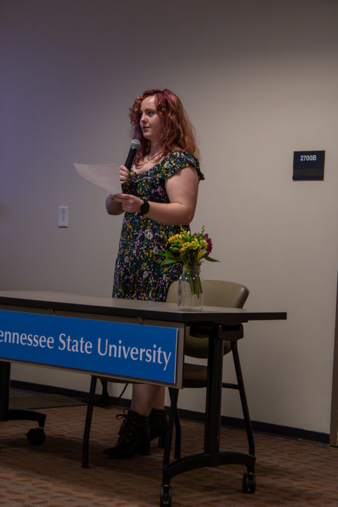 A student stands behind a table reading poetry into a microphone, with a small bouquet of flowers on the table and an MTSU sign displayed.